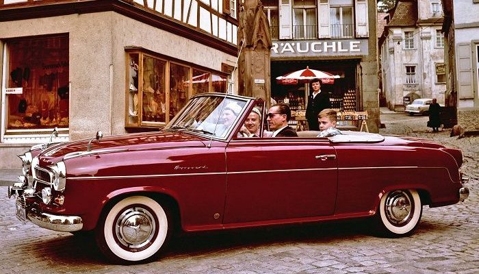 Ein weinrotes Borgward Cabriolet fährt über einen alten Stadtplatz