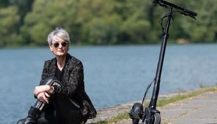 Frau sitzt am Wasser, neben ihr steht ein E-Scooter