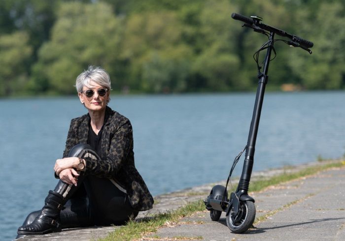 Frau sitzt am Wasser, neben ihr steht ein E-Scooter