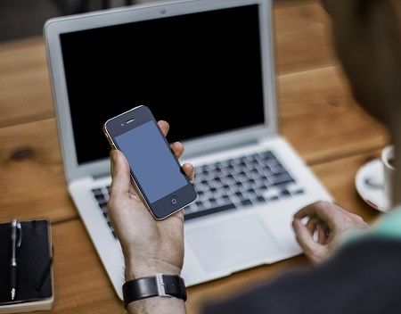 Mann sitzt mit einem Handy in der Hand vor seinem Laptop