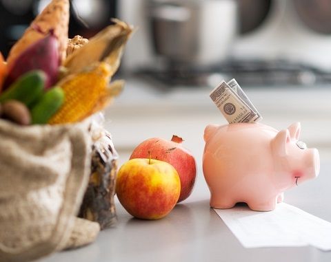 Sparschwein mit einem Geldschein, steht vor einer Einkaufstüte mit Gemüse