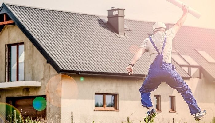 Mann springt vor einem renovierungsbedürftigem Haus, mit einem Plan, freudig in die Luft