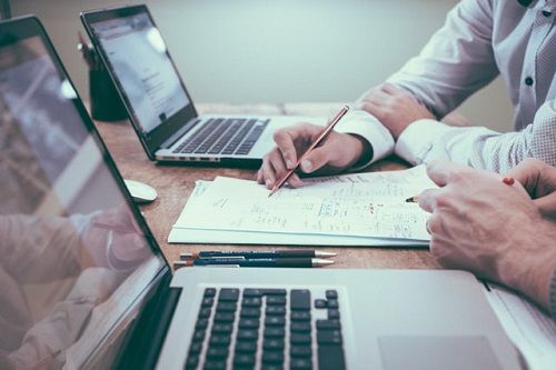 Zwei Männer sitzen vor Laptops und berechnen verschiedenes auf einem beigelegten Block