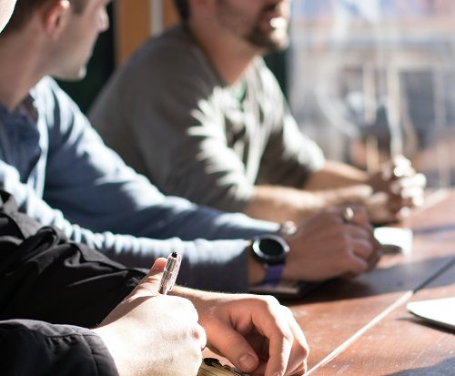 Männer sitzen, besprechend und mit Notizblöcken vor sich, an einem großen Besprechungstisch 