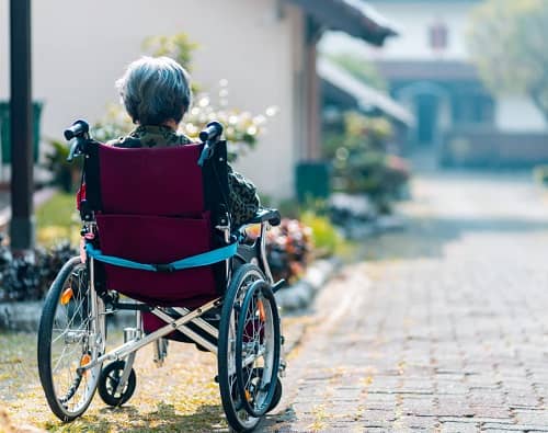 Frau sitzt im Rollstuhl und sieht über eine lange Einfahrt zu Ihrem Hauseingang