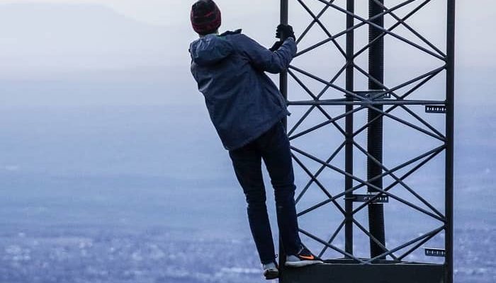 Mann steht hoch auf einem Strommast und blickt übers Meer auf eine gegenüber liegende Landschaft