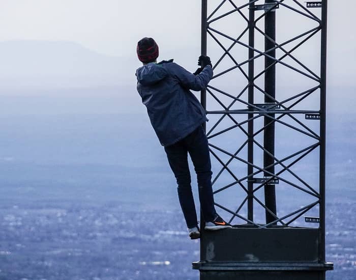 Mann steht hoch auf einem Strommast und blickt übers Meer auf eine gegenüber liegende Landschaft