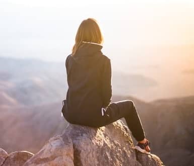 Frau sitzt auf einem Berggipfel und blickt in die Ferne