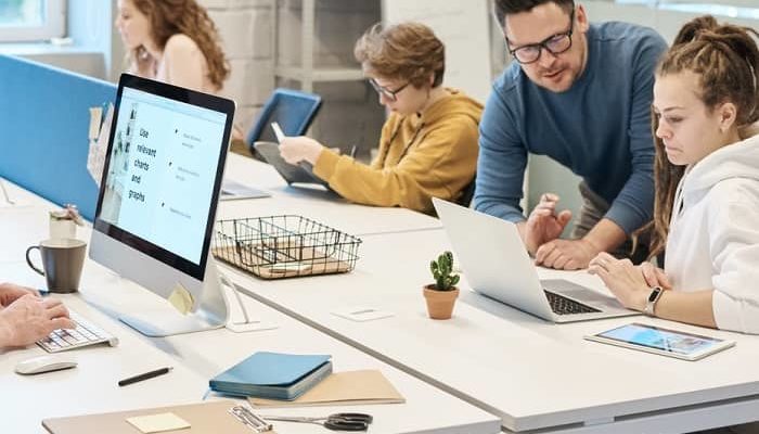 Ein Mann spricht mit einer Frau, in einem Büro wo mehrere Personen an Bildschirmen sitzen