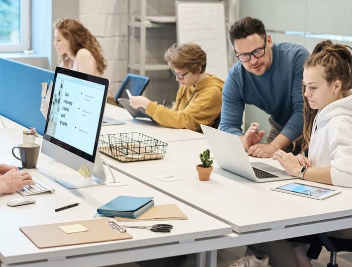 Ein Mann spricht mit einer Frau, in einem Büro wo mehrere Personen an Bildschirmen sitzen