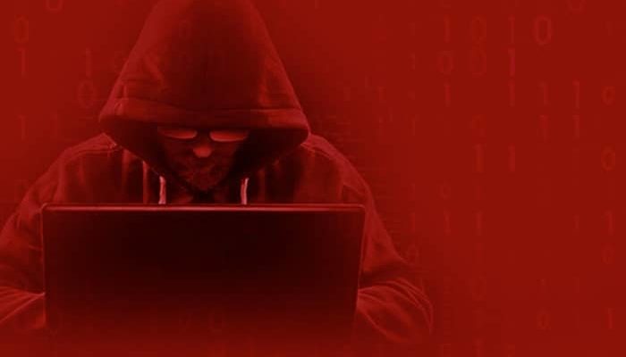 Hacker sitzt mit Laptop und vermummt, vor rotem Hintergrund und diversen Zahlenreihen