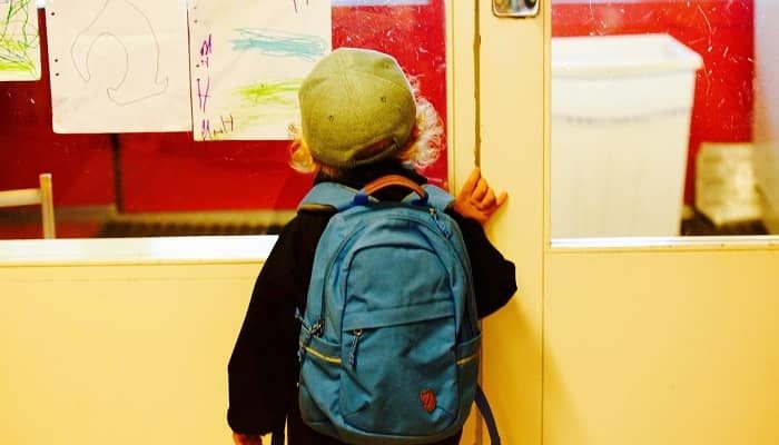 Jung steht vor einer Klassenzimmertür mit einem blauen Rucksack als Schulranzen am Rücken