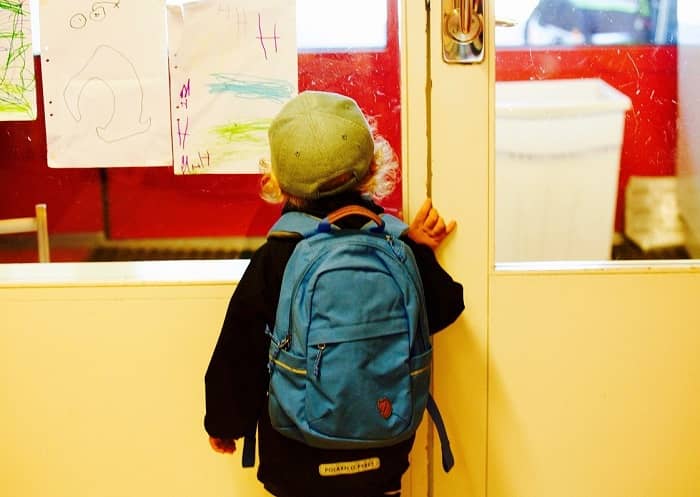 Jung steht vor einer Klassenzimmertür mit einem blauen Rucksack als Schulranzen am Rücken