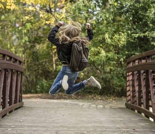 Mädchen springt auf einer Brücke im Park, mit Ihrem Schulranzen am Rücken, vor Freude in die Luft