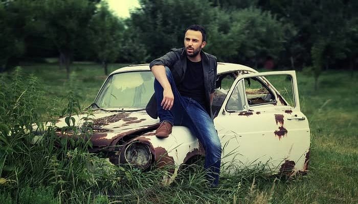 Ein Mann sitzt auf einem Schrottauto in mitten grüner Natur