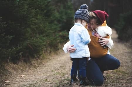 Mutter mit einem Baby vorne in der Bauchtragetasche spricht mit einem älteren Kind