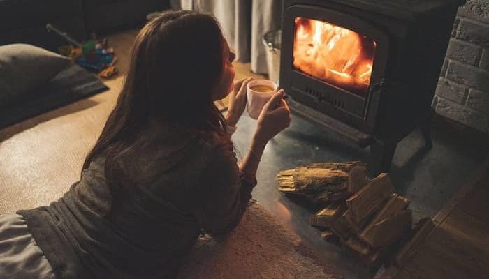 Frau liegt gemütlich mit einer Tasse Glühwein vor einem Ofen mit offenem Feuer