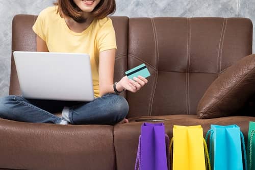 Frau sitzt mit einem Laptop und einer Kreditkarte auf der Couch, neben Ihr stehen Geschenktüten 