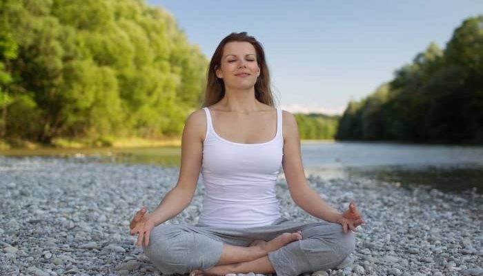 Frau sitzt beim Yoga im Schneidersitz auf einem Kiesstrand an einem Fluss