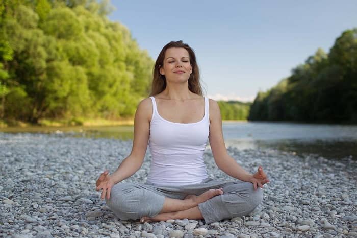 Frau sitzt beim Yoga im Schneidersitz auf einem Kiesstrand an einem Fluss