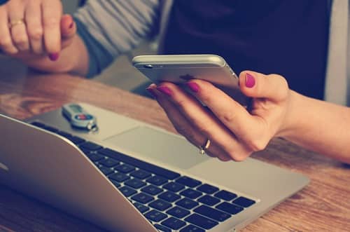 Eine Frau sitzt mit Ihrem Smartphone in der Hand vor Ihrem Laptop