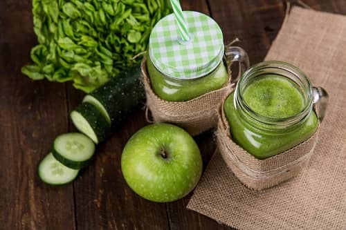 Zwei Gläser mit grünen Smoothies und ein Apfel und eine Gurke befinden sich auf einem Tisch