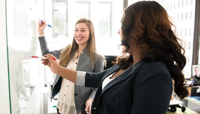 Zwei Frauen mit guter Laune stehen mit Stiften vor einem Whiteboard