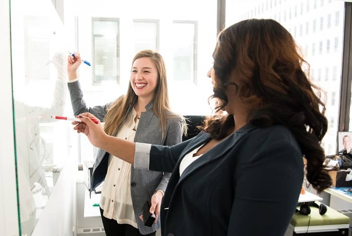 Zwei Frauen mit guter Laune stehen mit Stiften vor einem Whiteboard