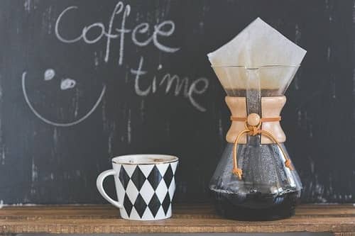 Eine Tasse Kaffee steht neben einer Kaffeekanne auf der gerade Kaffee handgebrüht wird