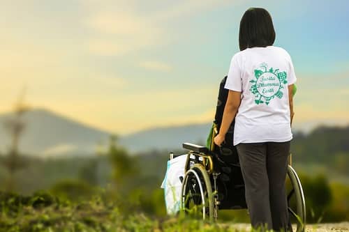 Eine Pflegerin schiebt einen Rollstuhl, im Hintergrund Berge und Natur