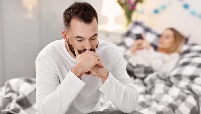 Ein Mann sitzt frustriert auf dem Bett, hinter Ihm liegt seine Frau mit einem Handy in der Hand