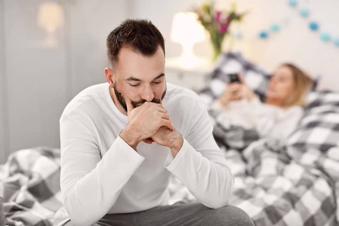 Ein Mann sitzt frustriert auf dem Bett, hinter Ihm liegt seine Frau mit einem Handy in der Hand