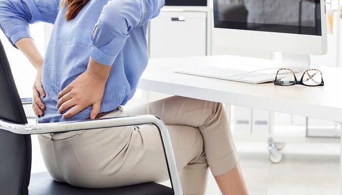 Eine Frau sitzt im Büro auf einem Stuhl und hält sich den unteren Rücken wegen Schmerzen