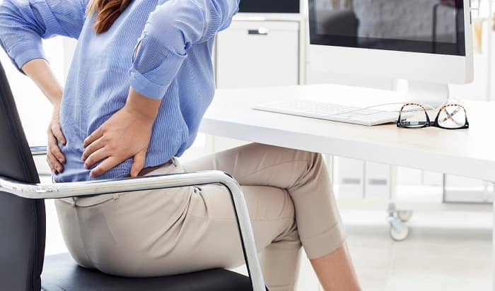 Eine Frau sitzt im Büro auf einem Stuhl und hält sich den unteren Rücken wegen Schmerzen
