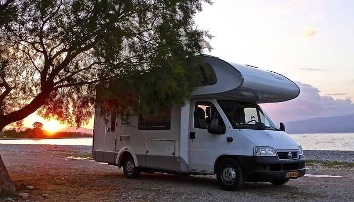 Ein Campingmobil steht im Sonnenuntergang auf einem Strand unter einem Baum.