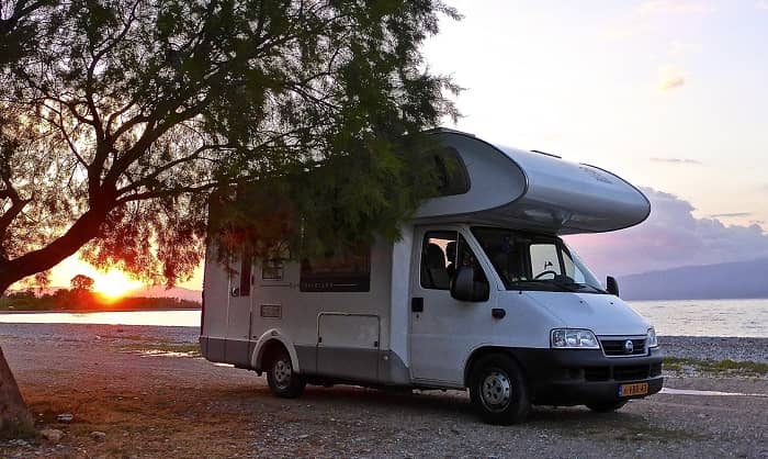 Ein Campingmobil steht im Sonnenuntergang auf einem Strand unter einem Baum.