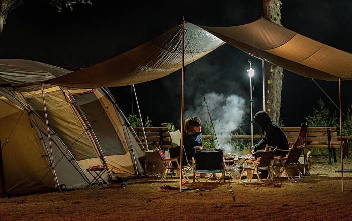 Ein großes Zelt unter dessen Vordach, nachts, zwei Personen grillen.