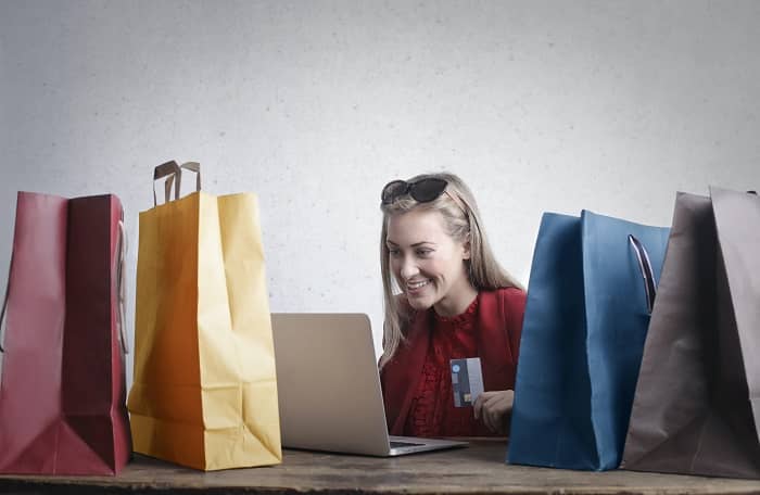 Eine Frau sitzt mit Laptop und Kreditkarte, umringt von Einkaufstüten, am Schreibtisch