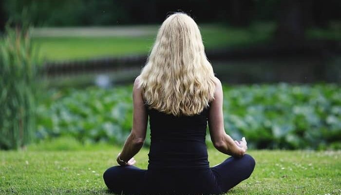 Eine Frau mit langen blonden Haaren sitzt vor einem Teich und meditiert