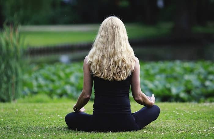 Eine Frau mit langen blonden Haaren sitzt vor einem Teich und meditiert
