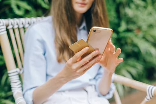 Eine Frau sitzt im Garten und bestellt etwas am Smartphone, Sie hat noch eine Kreditkarte in der Hand